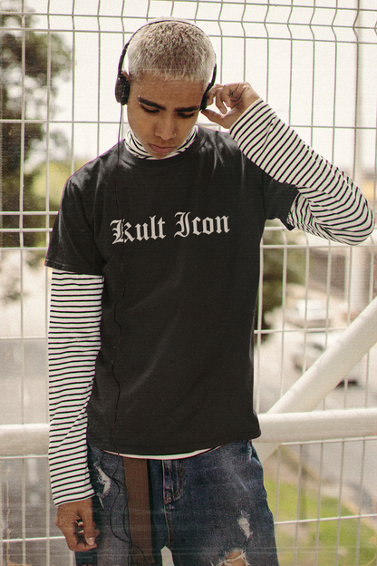 Person wearing a black 'Kult Icon' t-shirt with a striped long-sleeve shirt underneath, standing against a wire fence.