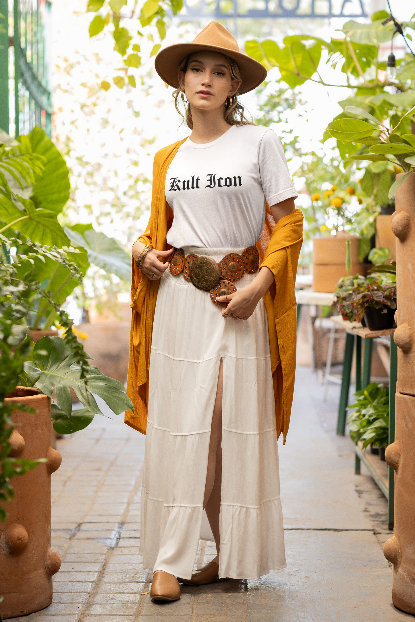Woman in a white outfit with a yellow cardigan and hat, standing in a garden.