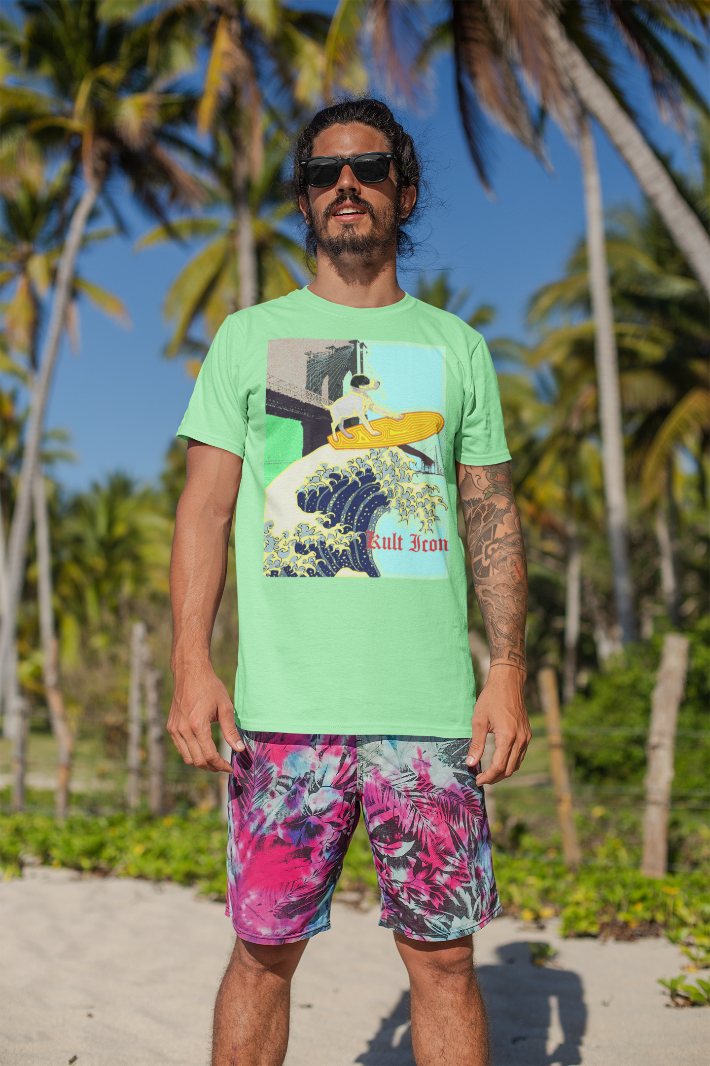 Man wearing a green t-shirt with a graphic design and colorful shorts on a beach with palm trees.
