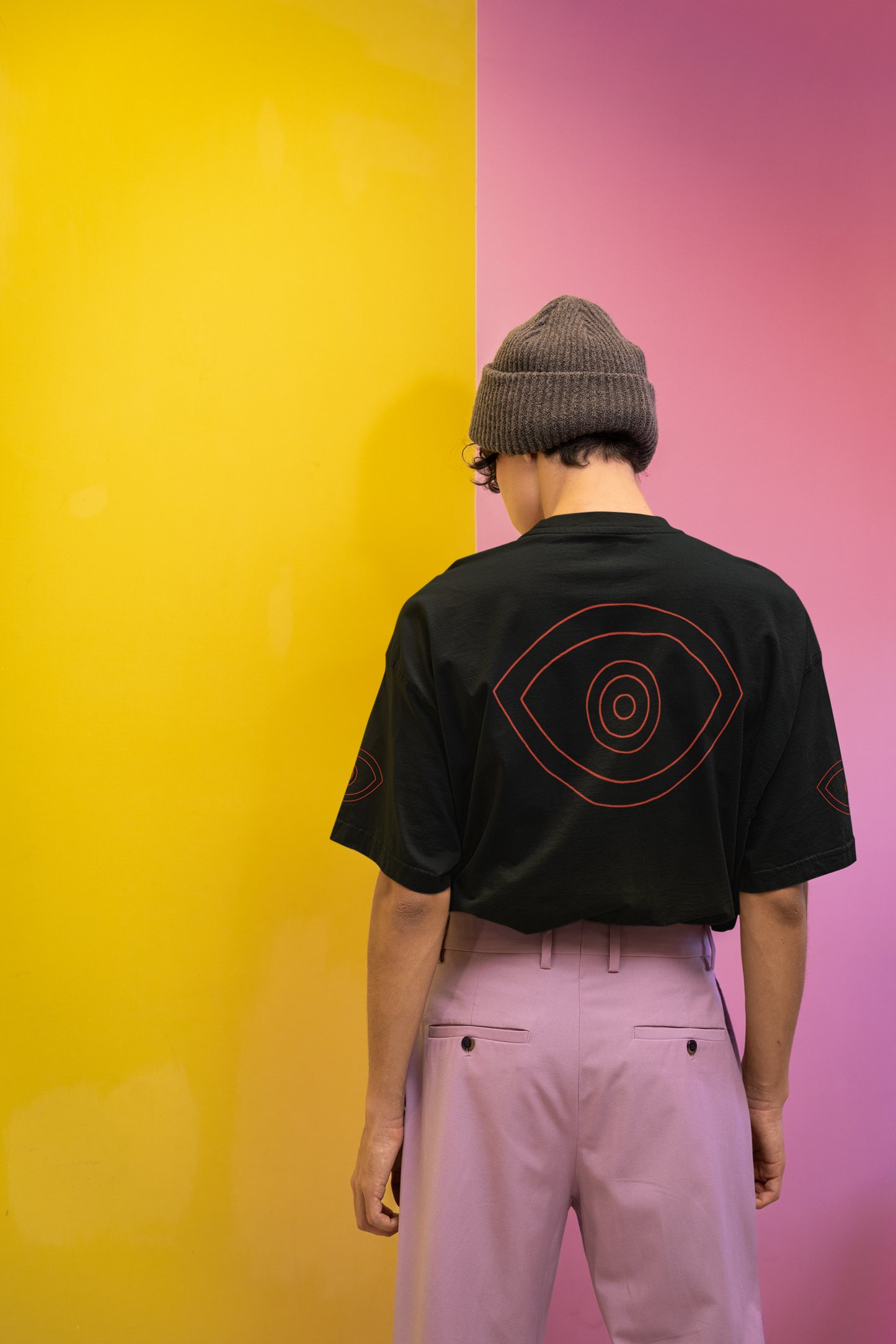 Person wearing a black t-shirt with a red eye design, standing against a yellow and pink split wall.